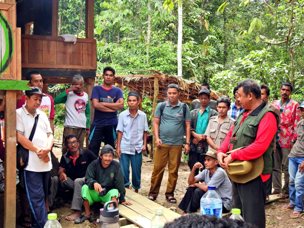 A man speaks to a group of participants standing and sitting in a forest area near a field post.