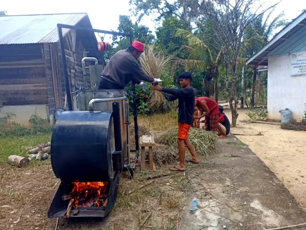 Several people loading plant material into a metal distillation unit heated by a wood fire.