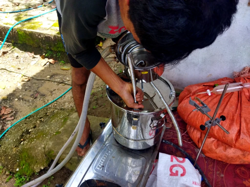 A participant adjusting hoses and metal components on a small-scale distillation setup outdoors.