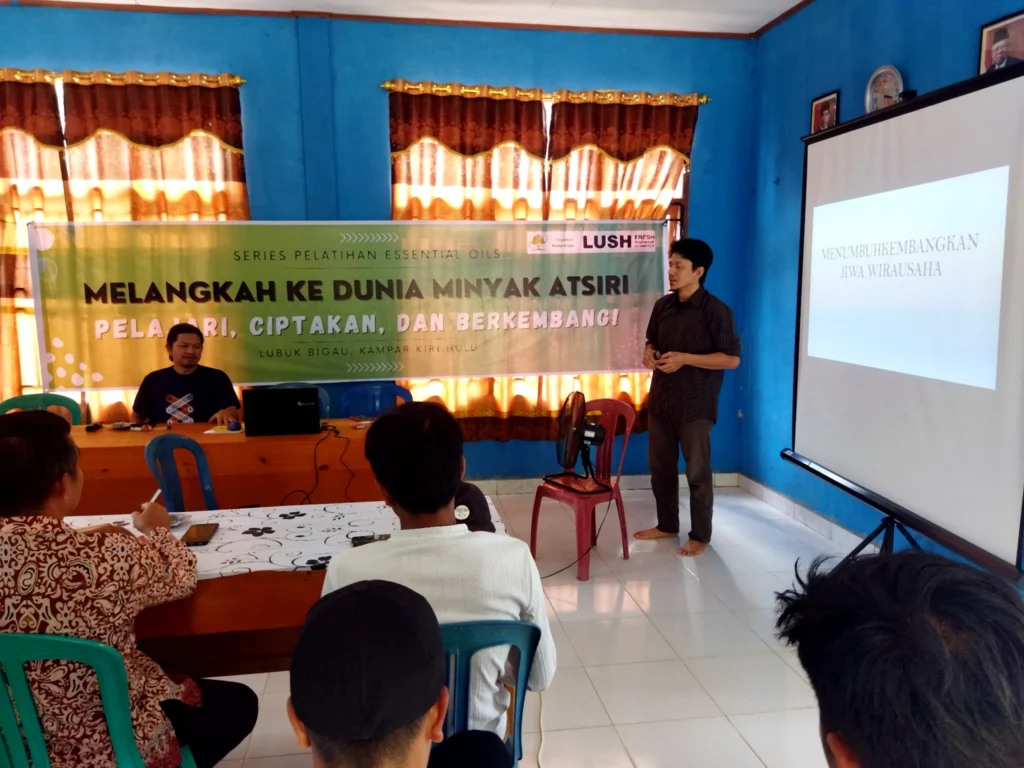 A facilitator presenting entrepreneurship material beside an essential-oil training banner and projection screen.