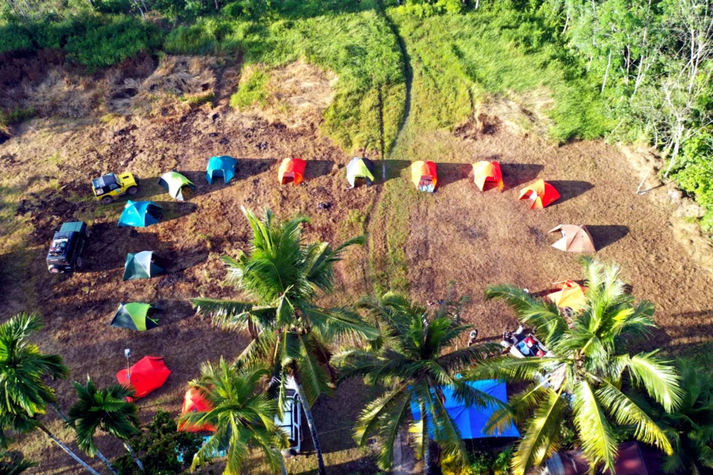 An aerial view of colourful tents and vehicles at a campsite bordered by palms and vegetation.