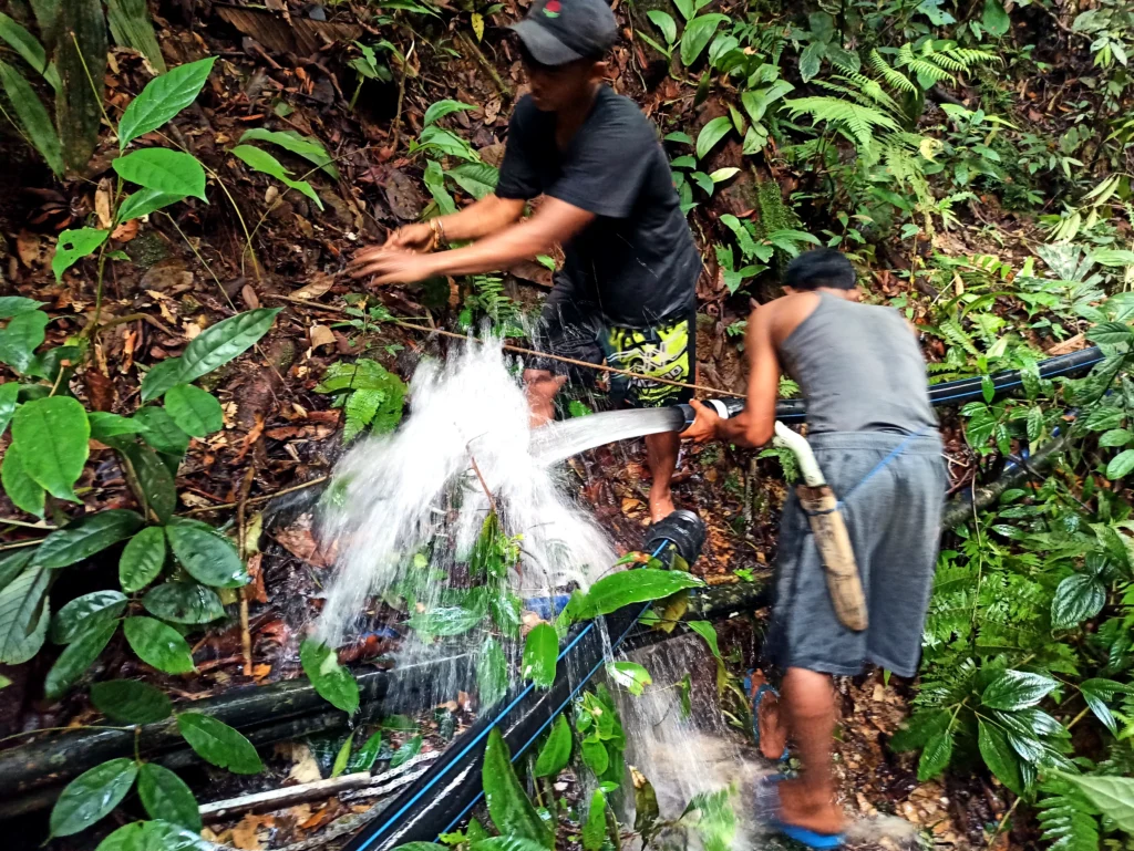 Two people handling pipes and materials beside a forest stream in Lubuk Bigau.