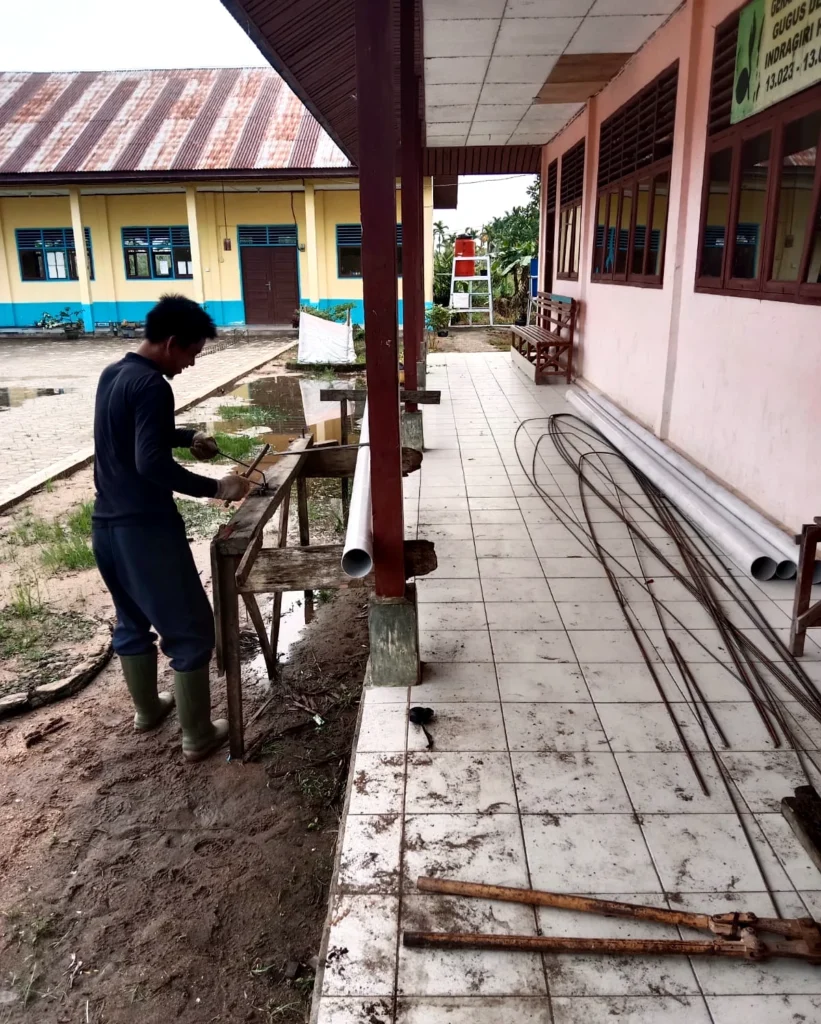 A worker cutting material along a school corridor with pipes and reinforcement bars nearby.
